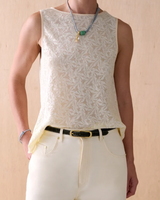A person wearing a sleeveless cream lace top with a leaf pattern, paired with cream pants and a olive green croc belt, standing against a light wood background.