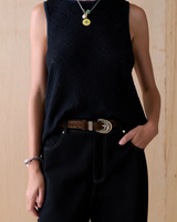 A model wearing a sleeveless black textured knit top and black wide-leg trousers, accessorized with a brown leather belt with a gold and silver buckle, a green and yellow pendant necklace, and a silver bracelet, set against a light wood panel background.